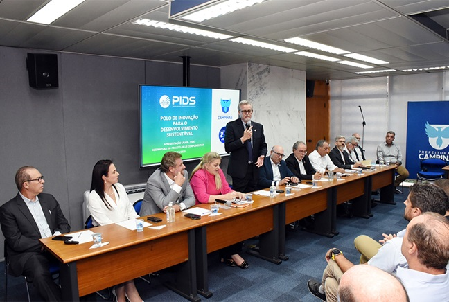 imagem de um grupo de pessoas sentadas lado a lado em uma sala de reuniões. Um dos homens está em pé e segura um microfone